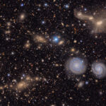 A sprawling, textured field of galaxies scattered across the deep black of space. It is filled with the delicate smudges and glowing cores of galaxies of many shapes, sizes and colors, as well as the bright multi-colored points of stars. To the lower left is a region filled with the hundreds of golden glittering gems of a distant galaxy cluster. In the foreground, below and right of center, two blue spiral galaxies look like eyes beneath the entangled mass of a triple galaxy merger in the upper right. A few bright blue points of foreground stars pierce the glittering tapestry. All throughout the image, thousands of galaxies gather in clusters or are spread throughout, like glittering gems strewn on a table. Some are sharp-edged and spiral, like coiled ribbons; others round and diffuse, like polished pebbles. Still others are just smudges of various colors against the black of space. The background is peppered with pinpoint stars in reds, yellows, and blues, crisp against the velvet black.