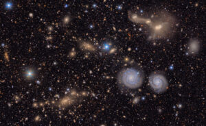 A sprawling, textured field of galaxies scattered across the deep black of space. It is filled with the delicate smudges and glowing cores of galaxies of many shapes, sizes and colors, as well as the bright multi-colored points of stars. To the lower left is a region filled with the hundreds of golden glittering gems of a distant galaxy cluster. In the foreground, below and right of center, two blue spiral galaxies look like eyes beneath the entangled mass of a triple galaxy merger in the upper right. A few bright blue points of foreground stars pierce the glittering tapestry.

All throughout the image, thousands of galaxies gather in clusters or are spread throughout, like glittering gems strewn on a table. Some are sharp-edged and spiral, like coiled ribbons; others round and diffuse, like polished pebbles. Still others are just smudges of various colors against the black of space. The background is peppered with pinpoint stars in reds, yellows, and blues, crisp against the velvet black.